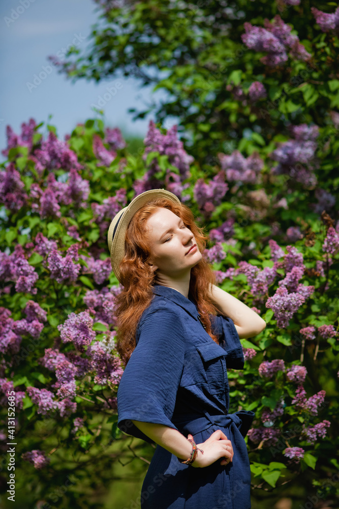 A young woman enjoys the smell of lilac on a sunny summer day. Aromatherapy and the concept of spring. A beautiful woman in a blue long vintage dress and a straw hat stands in a lilac garden. A young woman enjoys the smell of lilac on a sunny summer day. Aromatherapy and the concept of spring. A beautiful woman in a blue long vintage dress and a straw hat stands in a lilac garden.