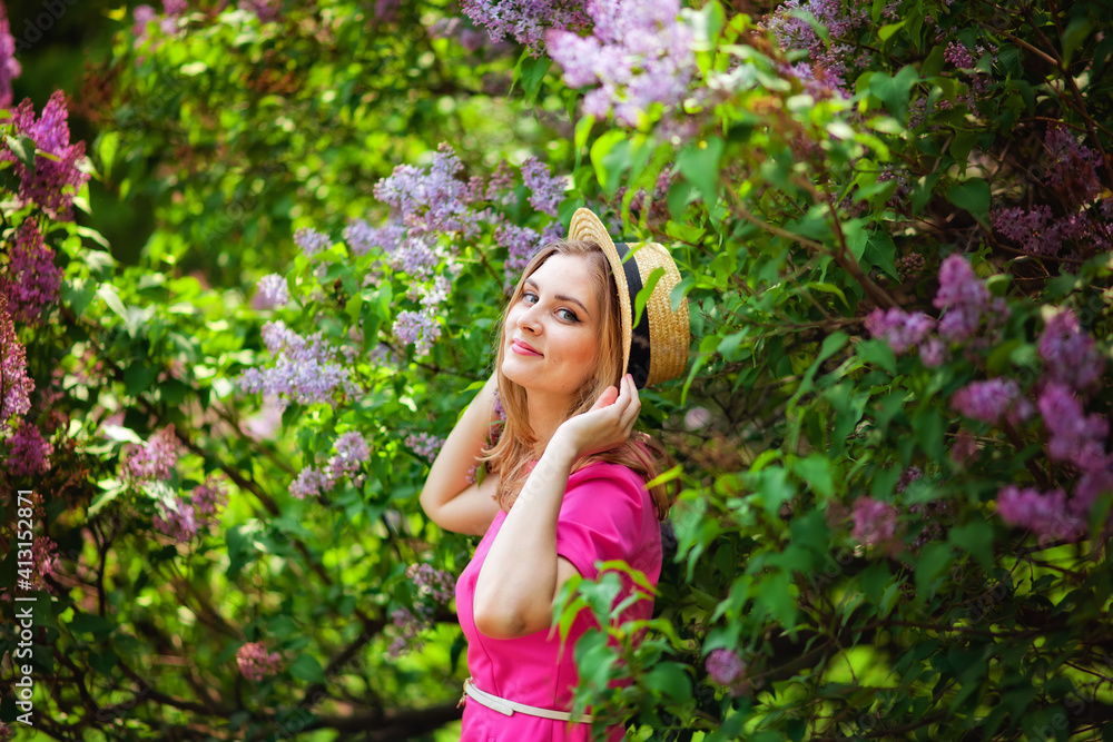 A young woman enjoys the smell of lilac on a sunny summer day. Aromatherapy and the concept of spring. A beautiful woman in a pink dress and a straw hat stands in a lilac garden. A young woman enjoys the smell of lilac on a sunny summer day. Aromatherapy and the concept of spring. A beautiful woman in a pink dress and a straw hat stands in a lilac garden.