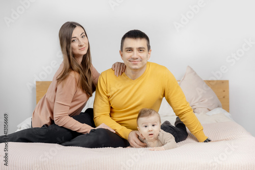 Happy young family woman, man and child are sitting on bed and smiling, light background