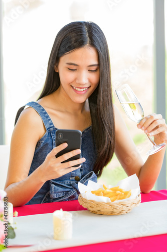 A cute beautiful young Asian woman sitting alone at the fast food shop and holding wine glass and smartphone