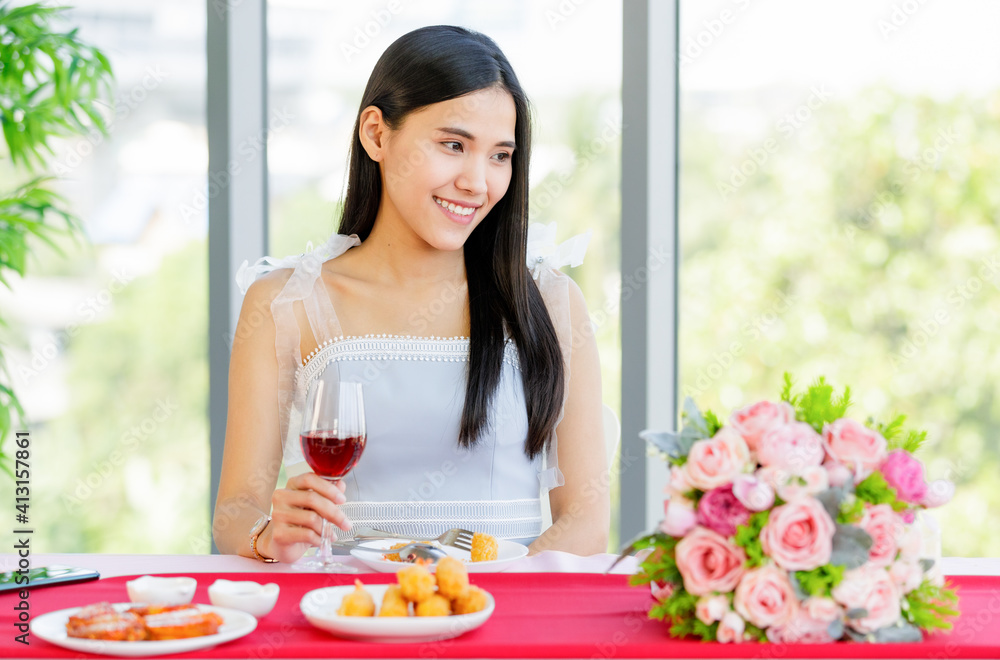 A beautiful young cute black long hair Asian woman sitting in the restaurant with a flower bouquet and holding a red wine glass in hand with a smile happy face