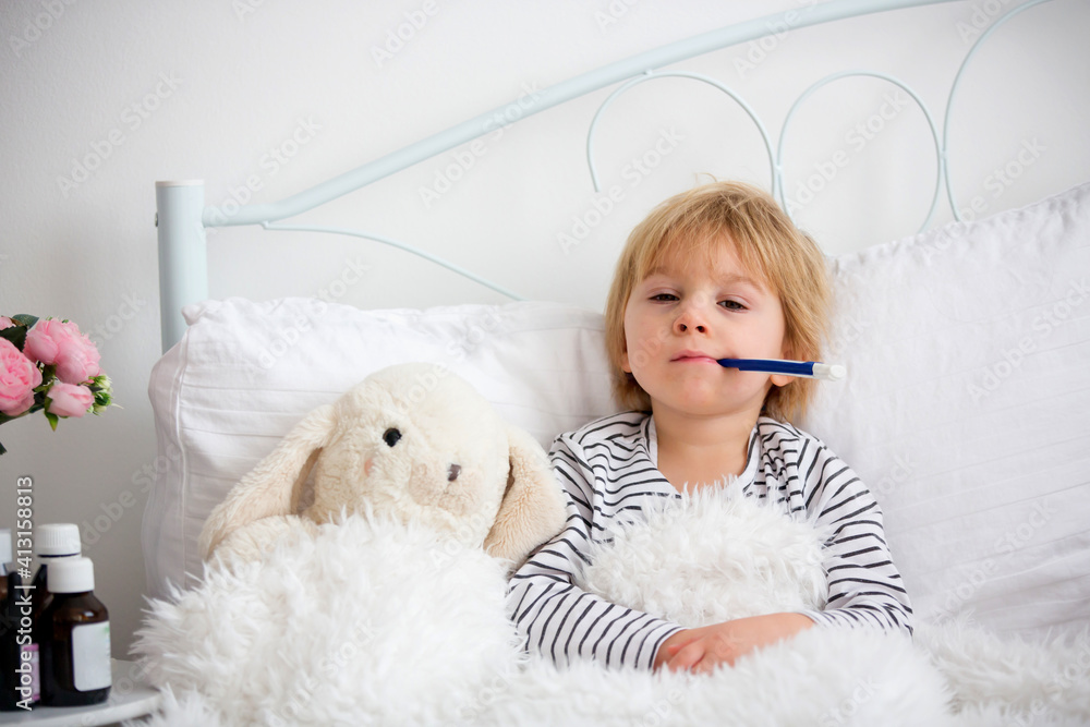 Sick little blond toddler boy, lying in bed with fever, hugging little fluffy toy, medicine next to him Sick little blond toddler boy, lying in bed with fever, hugging little fluffy toy, medicine next to him