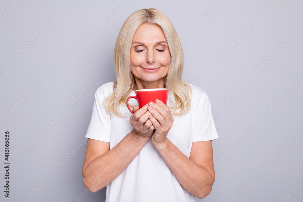 Photo portrait of old woman inhaling smell of nice morning coffee holding red cup in two hands isolated on grey colored background