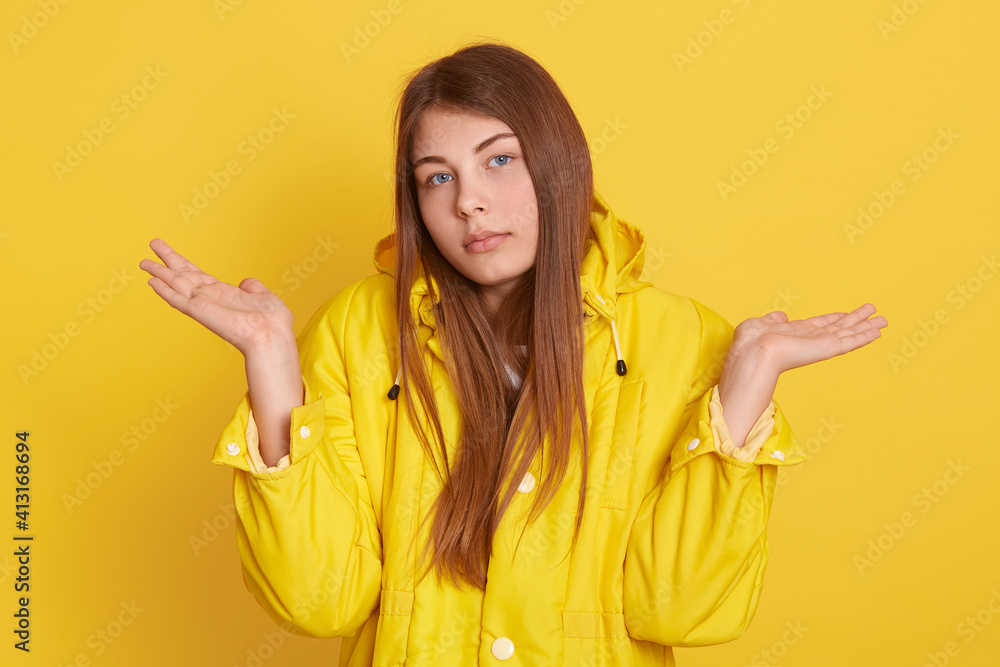 Clueless unaware woman with long hair, spreads palms in doubt, expresses uncertainty, has sad expression, wears yellow jacket, shrugs shoulders, looks at camera, isolated over color background. Clueless unaware woman with long hair, spreads palms in doubt, expresses uncertainty, has sad expression, wears yellow jacket, shrugs shoulders, looks at camera, isolated over color background.