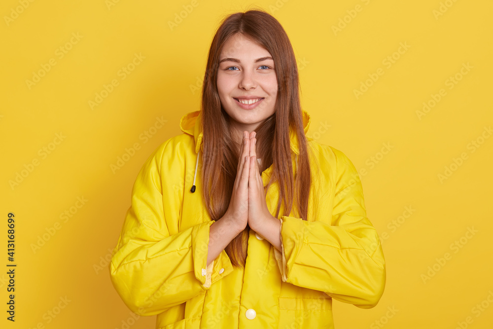 Joyous European girl looks with happy expression at camera, keeps hands pressed together in front of chest, has pleasant news, has natural pleasant appearance, poses indoor against yellow wall. Joyous European girl looks with happy expression at camera, keeps hands pressed together in front of chest, has pleasant news, has natural pleasant appearance, poses indoor against yellow wall.