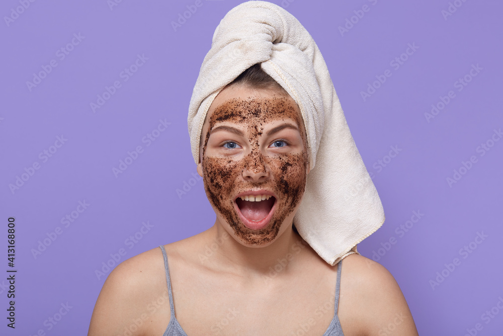 Exited beautiful girl wearing sleeveless t shirt and wrapped towel, doing cosmetic procedures for peeling skin, woman yelling something happily isolated over lilac wall, cleansing face skin via scrub. Exited beautiful girl wearing sleeveless t shirt and wrapped towel, doing cosmetic procedures for peeling skin, woman yelling something happily isolated over lilac wall, cleansing face skin via scrub.