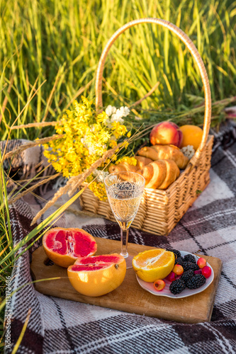 Picnic basket with food and glass with champagne on green sunny lawn. there are two croissants, grapefruit, orange, berries, peaches. photo