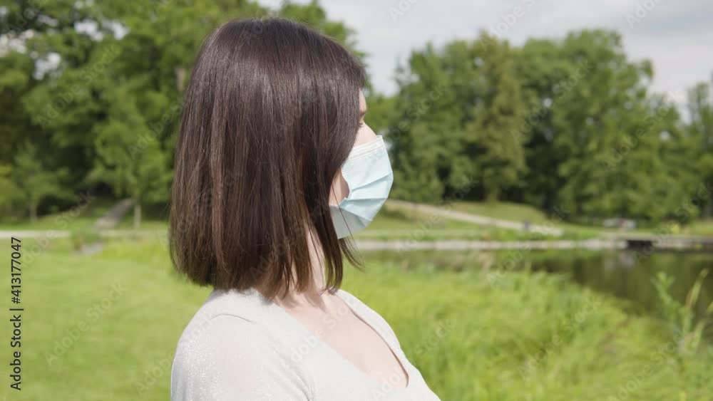 A young Caucasian woman in a face mask looks around thoughtfully by a pond in a city park on a sunny day - trees and a pathway in the blurry background A young Caucasian woman in a face mask looks around thoughtfully by a pond in a city park on a sunny day - trees and a pathway in the blurry background
