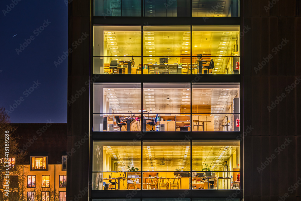 A contemporary office with a broad modern, glazed facade illuminated at night. Square pattern of wide glazed windows of a financial firm exterior. Tech industry headquarter with office space interiors - obrazy, fototapety, plakaty 