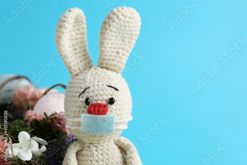 Toy bunny in protective mask on light blue background, space for text. Easter holiday during COVID-19 quarantine