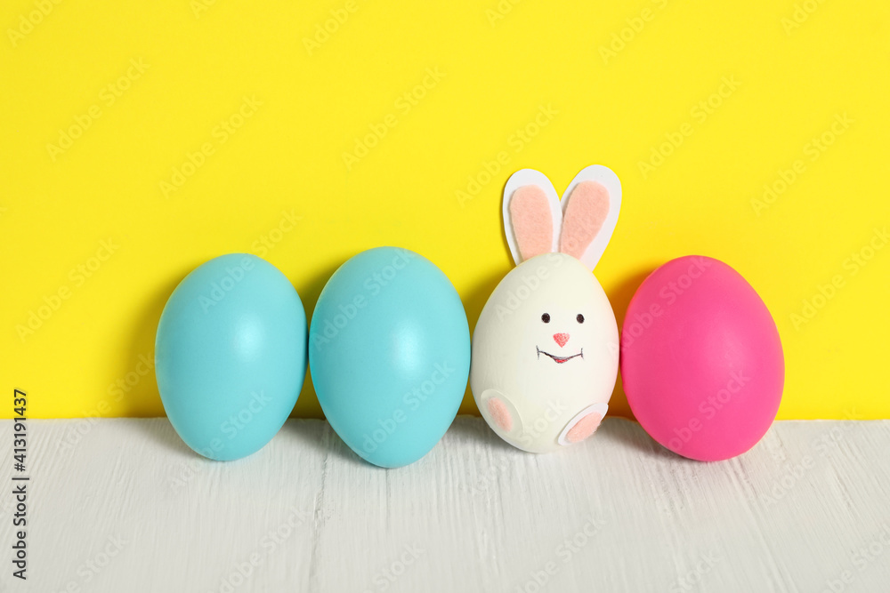 Bright Easter eggs and white one as cute bunny on wooden table against yellow background