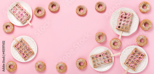 Traditional belgian waffles with sprinkles and delicious doughnuts with powdered sugar on pastel pink background.