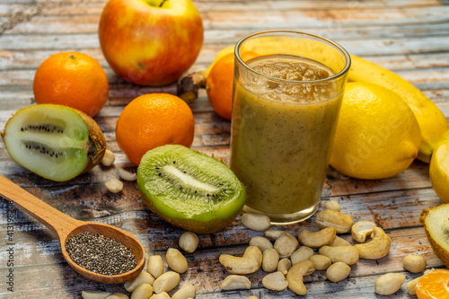 Green fresh healthy juice with fruits and vegetables on cutting board and wooden table background. Tangerines, kiwi, lemon, chia, nuts, banana and apple in the background. Selective focus