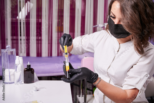Canvas Print Cosmetologist wearing black gloves, face mask and white uniform, sitting at the table, during process of filling syringe with plasma from a blood tube