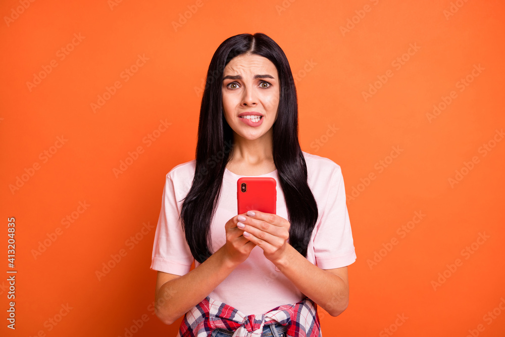 Portrait of young worried blogger biting lip hands hold phone look camera isolated on orange color background Portrait of young worried blogger biting lip hands hold phone look camera isolated on orange color background