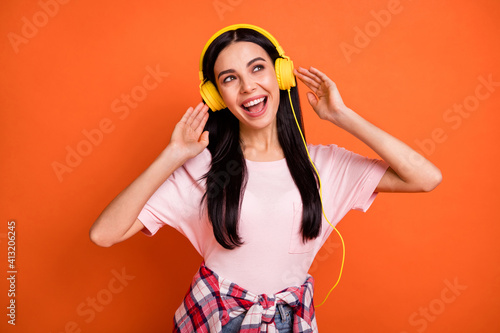 Photo of cheerful nice girl hands touch headphones look empty space isolated on orange color background