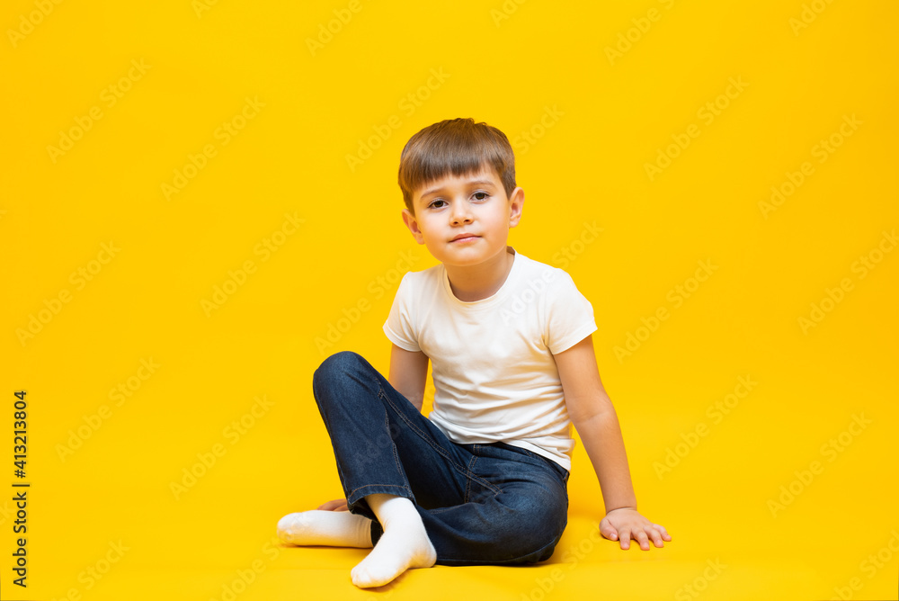 Naklejka premium Little boy in jeans and a white t-shirt sits on an isolated yellow background