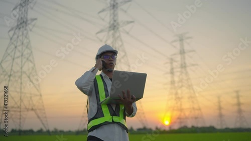 An electrical engineer with a laptop talking on the phone High voltage tower inspection report before starting a project Assigned by the organization during the time of sunset or sunrise.