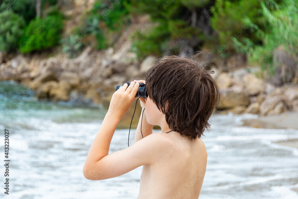 A young boy looking through binoculars staying at the seaside on the beach. Child spending summer holidays at the mountainous coast.