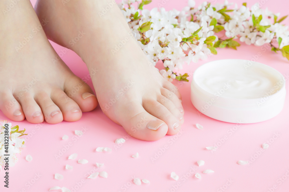 Young adult woman feet. Opened white jar of natural herbal cream on light pastel pink background. Care about clean, soft and smooth skin. Beautiful branch of cherry blossoms. Fresh flowers. Closeup. Young adult woman feet. Opened white jar of natural herbal cream on light pastel pink background. Care about clean, soft and smooth skin. Beautiful branch of cherry blossoms. Fresh flowers. Closeup.