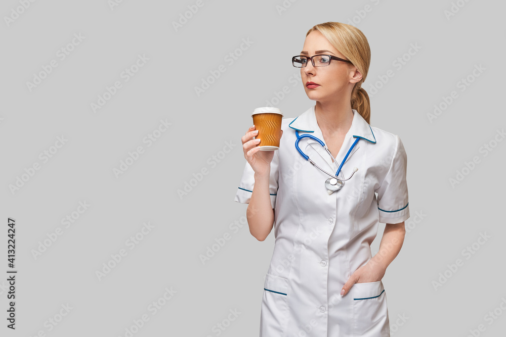 Portrait of a female doctor holding paper cup of coffee standing over grey background Portrait of a female doctor holding paper cup of coffee standing over grey background
