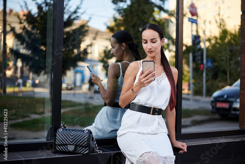 Young brunette girl with red pony tail, wearing white silk dress, sitting near modern glass building, holding phone. Pretty business woman on lunch break. Female city portrait