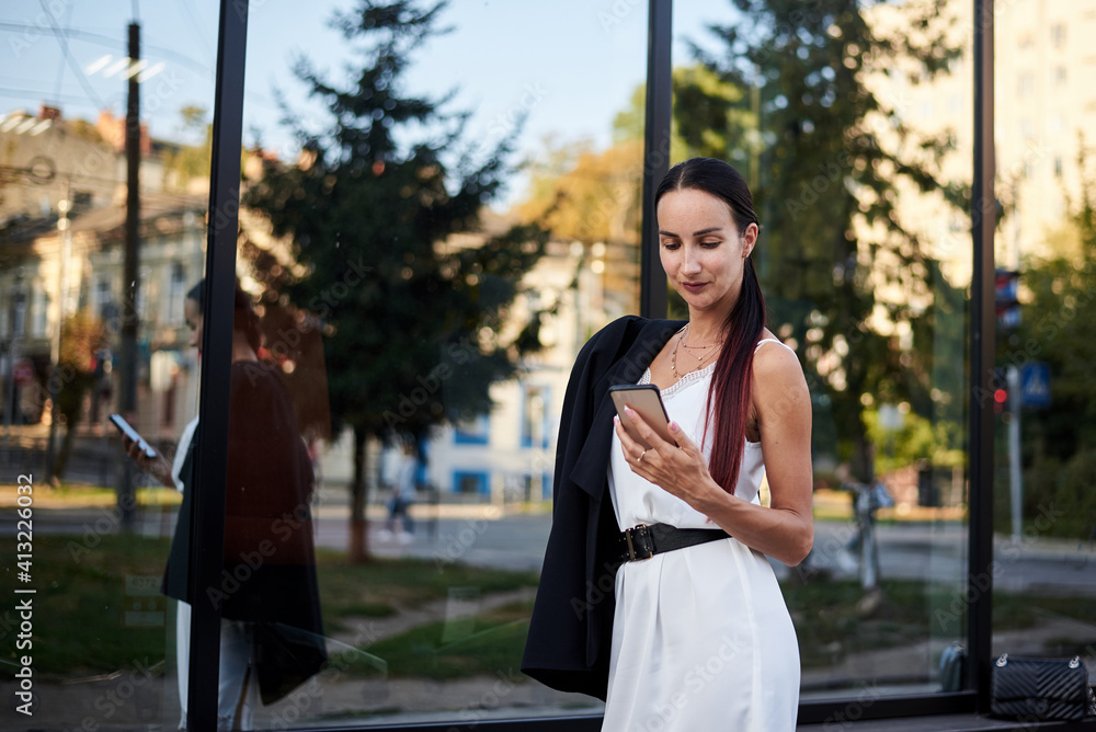 Young brunette girl with red pony tail, wearing stylish black jacket and white silk dress, standing near modern glass building, holding phone. Pretty business woman on lunch break. Female portrait