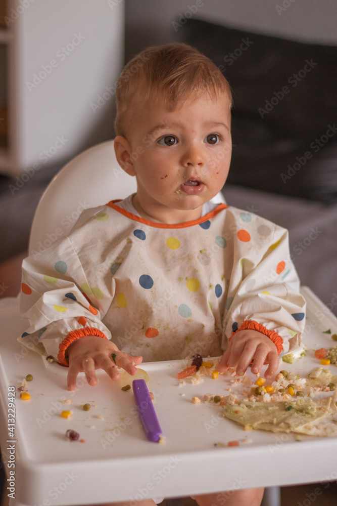 Baby boy sitting in highchair and eating healthy lunch. Baby learning to eat has food on his face and colorful clothes with surprised face expression. Baby boy sitting in highchair and eating healthy lunch. Baby learning to eat has food on his face and colorful clothes with surprised face expression.