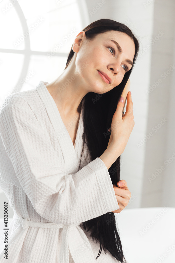 Young brunette woman combing her healthy and shiny hair sitting on bathtub weared in white bathrobe Young brunette woman combing her healthy and shiny hair sitting on bathtub weared in white bathrobe