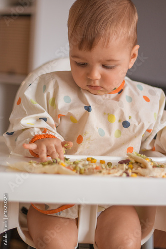Baby boy sitting in highchair and eating healthy lunch. Baby learning to eat has food on his face and colorful clothes. 