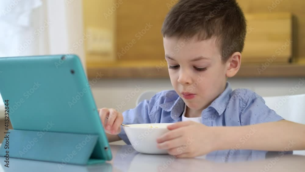 Little boy eating cereal with milk in the kitchen and watching cartoon on tablet computer.
