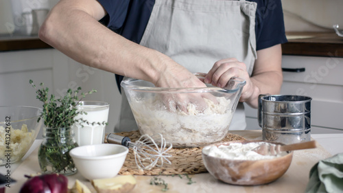 A faceless man in an apron in his kitchen at the kitchen table kneads bread dough in a glass bowl with his hands. Home authentic hobby, home baker. Baking bread with your own hands