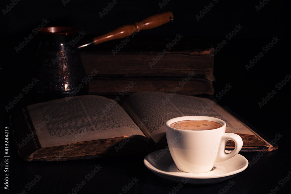 Cup of hot coffee, copper cezve and open Torah page on black wood table in the dark. Closeup.