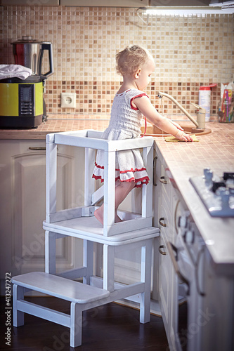 Little Girl Washing Dishes in the Kitchen. Stands on a folding Ladder-Table Chair.