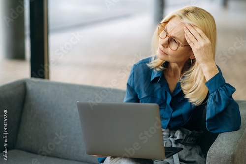 Senior adult business woman is sitting on the couch, using laptop. Exhausted female employee is having a terrible headache, feeling bad after tense brainstorm, overwork concept