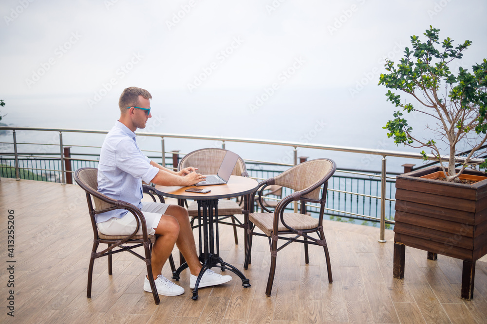Handsome successful young male businessman sitting at a table by the pool with a laptop overlooking the Mediterranean Sea. Remote work on vacation. Vacation concept