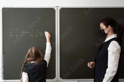 teacher and pupil in face pritection masks in front of blackboard solve math task in primary school - new normal concept photo