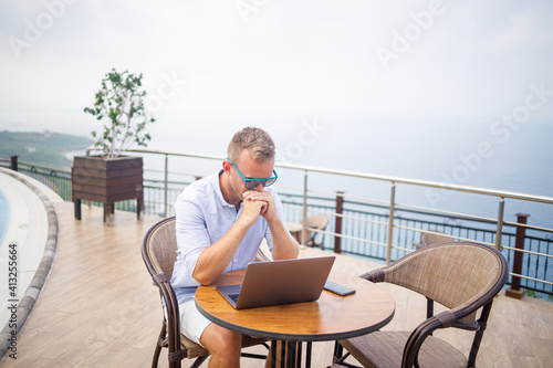 Handsome successful young male businessman sitting at a table by the pool with a laptop overlooking the Mediterranean Sea. Remote work on vacation. Vacation concept © Дмитрий Ткачук