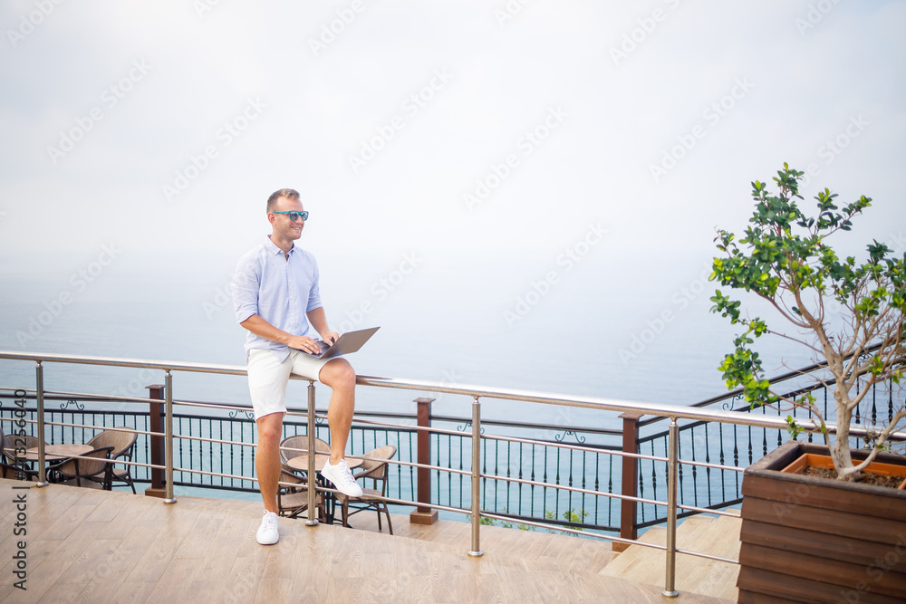 Handsome successful young male businessman working with laptop looks at the Mediterranean Sea. He is wearing a shirt and white shorts. Remote work on vacation. Vacation concept Handsome successful young male businessman working with laptop looks at the Mediterranean Sea. He is wearing a shirt and white shorts. Remote work on vacation. Vacation concept