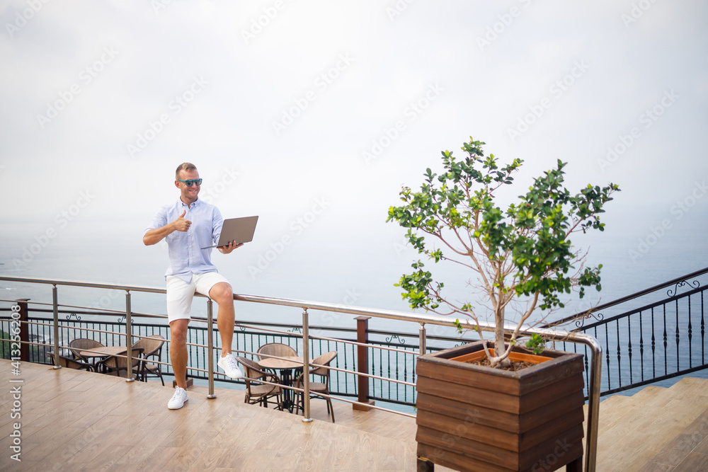 Handsome successful young male businessman working with laptop looks at the Mediterranean Sea. He is wearing a shirt and white shorts. Remote work on vacation. Vacation concept