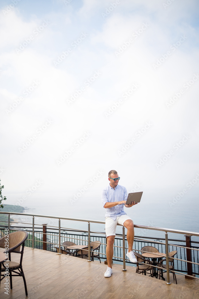 Handsome successful young male businessman working with laptop looks at the Mediterranean Sea. He is wearing a shirt and white shorts. Remote work on vacation. Vacation concept Handsome successful young male businessman working with laptop looks at the Mediterranean Sea. He is wearing a shirt and white shorts. Remote work on vacation. Vacation concept
