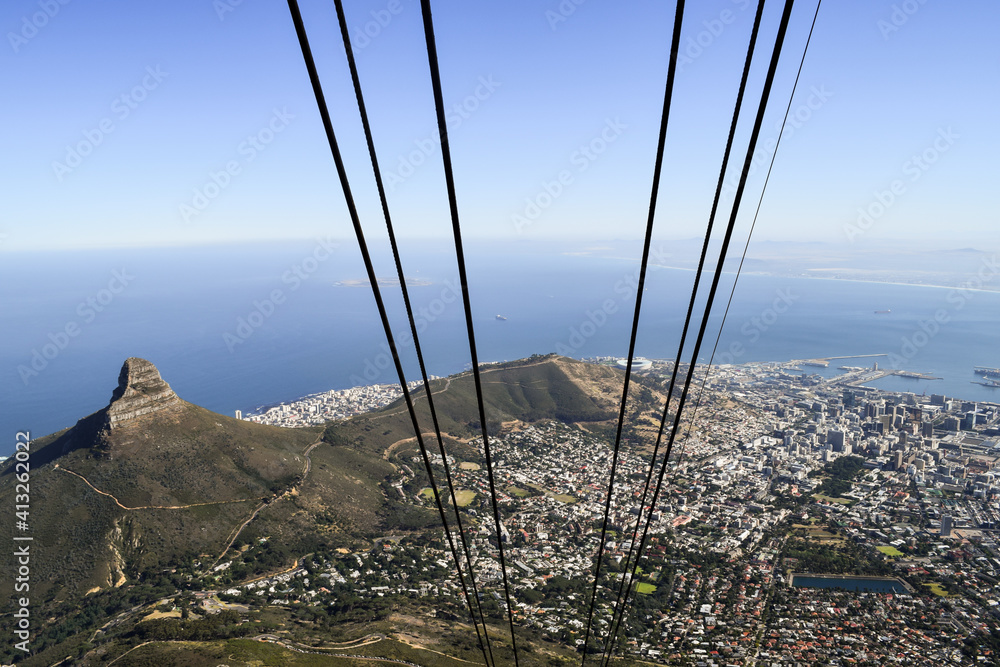 Fototapeta premium View of Cape Town from inside the cable car that takes people up to Table Mountain. The cables are in the foreground. The city, sea, Lion's Head in the background.