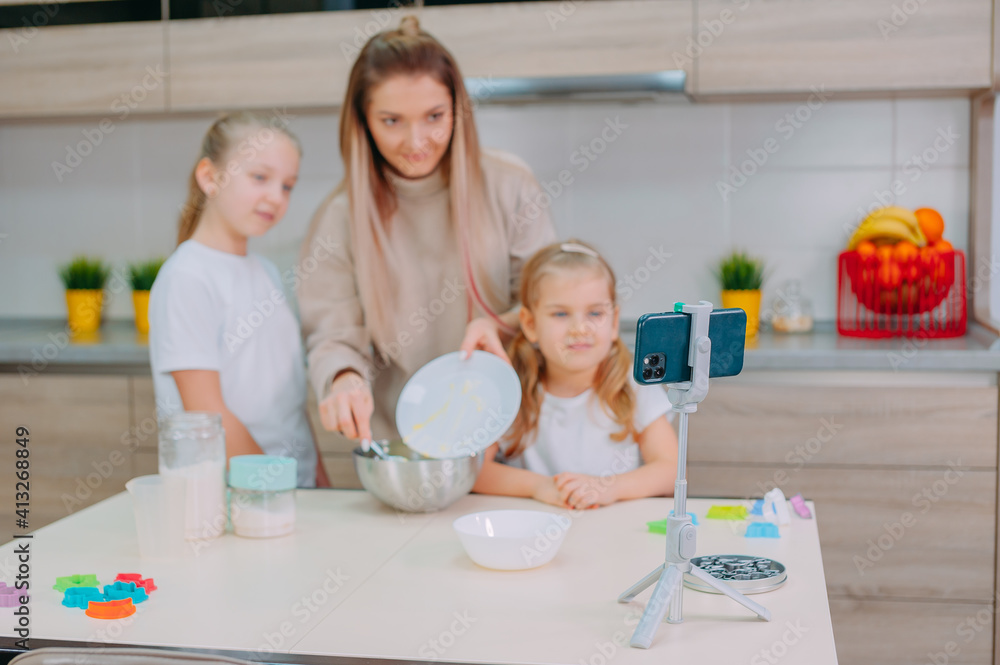 Fototapeta premium Mom teaches her daughters to cook dough in the kitchen. The family is filming a culinary video on a smartphone.