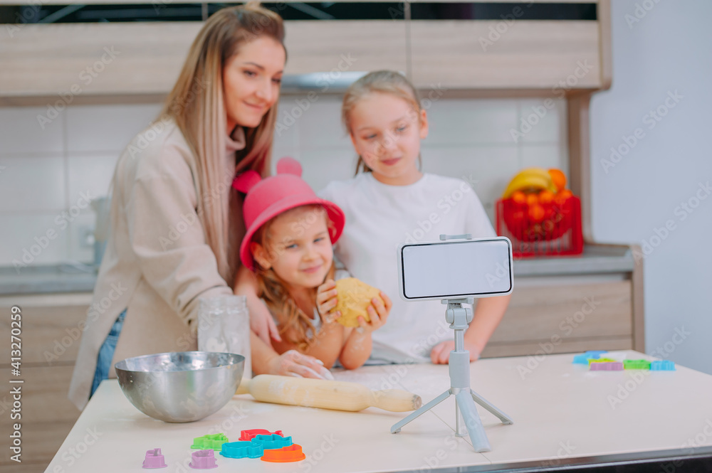 Fototapeta premium Mom teaches her daughters to cook dough in the kitchen. The family is filming a culinary video on a smartphone.