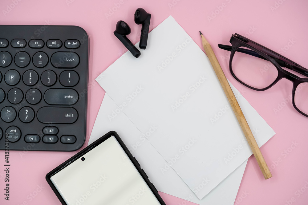 Top view of blank paper page on pink background office desk and different objects. Minimal flat lay style. With pencil, eye glasses, keyboard laptop, cellphone and ear phone Top view of blank paper page on pink background office desk and different objects. Minimal flat lay style. With pencil, eye glasses, keyboard laptop, cellphone and ear phone