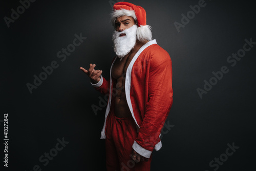 Surprising muscular man in Santa Claus costume in studio. Emotional portrait of handsome male model in Christmas outfit at grey background.