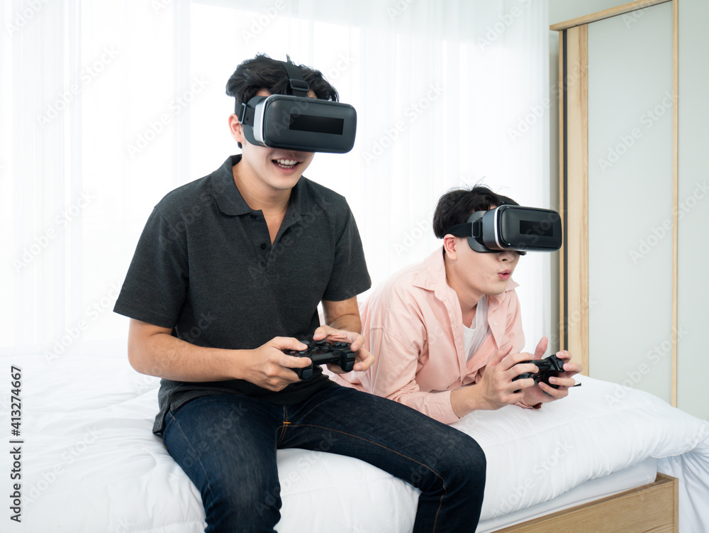 Asian young two boy immerse playing video game by virtual reality headset and joystick controller gadget on bed in white bedroom at comfort home Asian young two boy immerse playing video game by virtual reality headset and joystick controller gadget on bed in white bedroom at comfort home