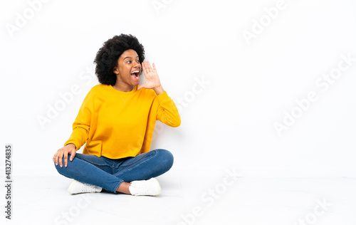 Young African American woman sitting on the floor shouting with mouth wide open to the lateral
