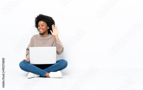 Young African American woman with a laptop sitting on the floor saluting with hand with happy expression © luismolinero