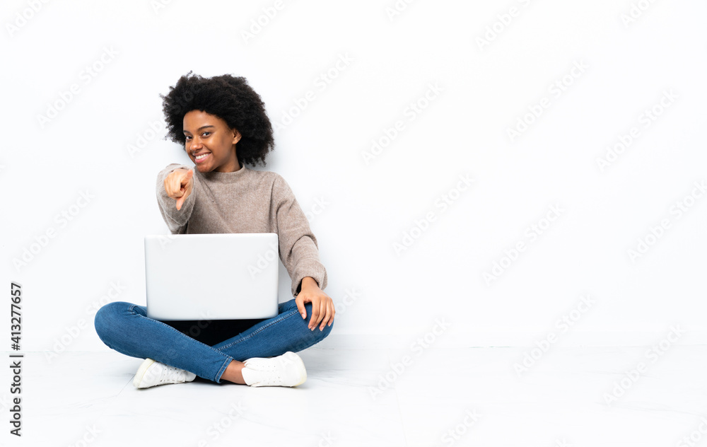 Young African American woman with a laptop sitting on the floor points finger at you with a confident expression